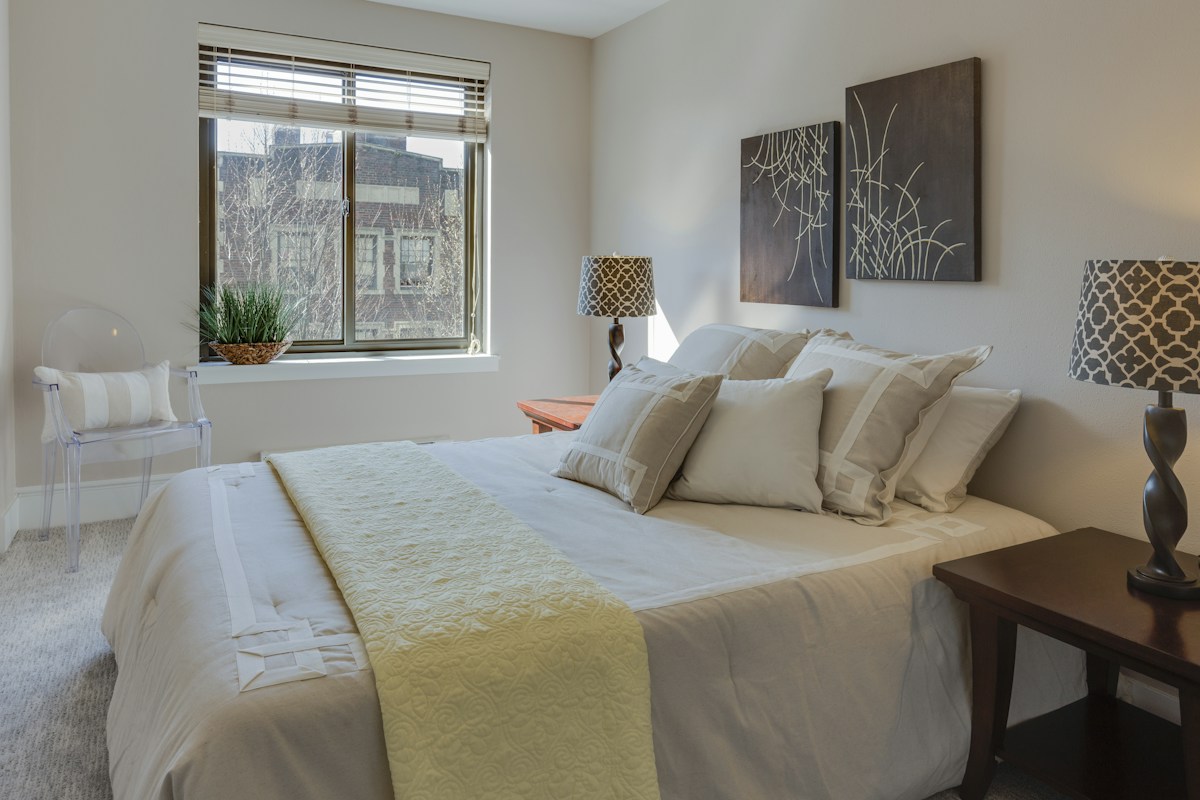 Layered bedding and textured cushions beside a window with venetian blinds